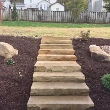 Wide natural stone steps leading up a landscaped slope with mulch, boulders, lawn, and a wooden fence.