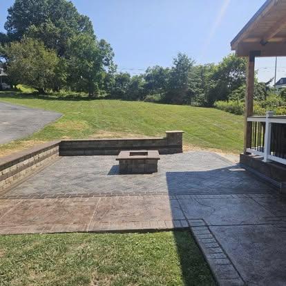 Stone patio with herringbone pavers, a square fire pit, low retaining wall, and open lawn views.