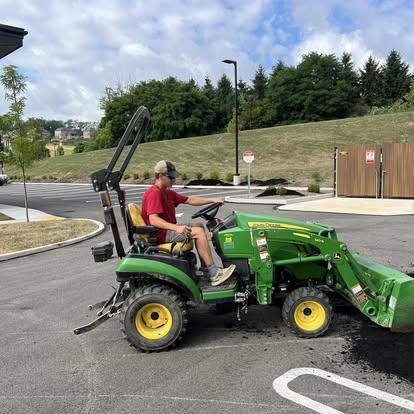 Operator using a John Deere compact tractor with front loader on an asphalt driveway.
