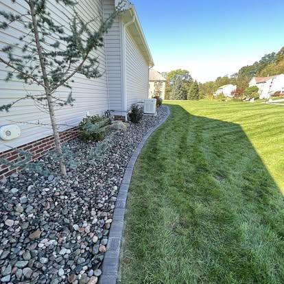Curved paver edging separating a river-rock bed from a green lawn along a home exterior.