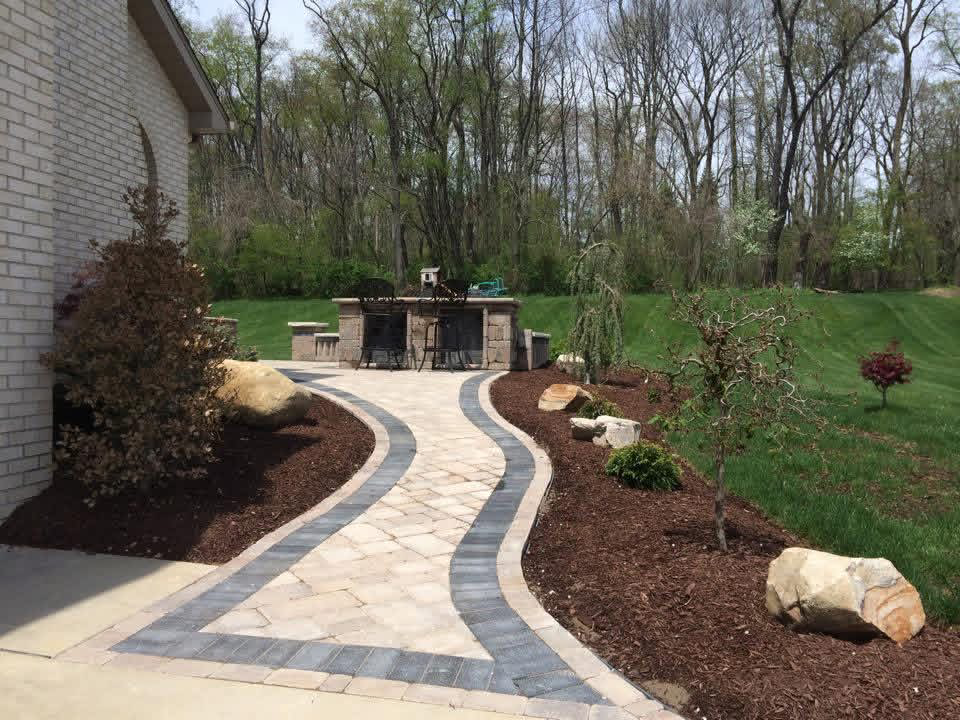 Curving paver walkway with dark border passing through mulched beds toward an outdoor stone bar.