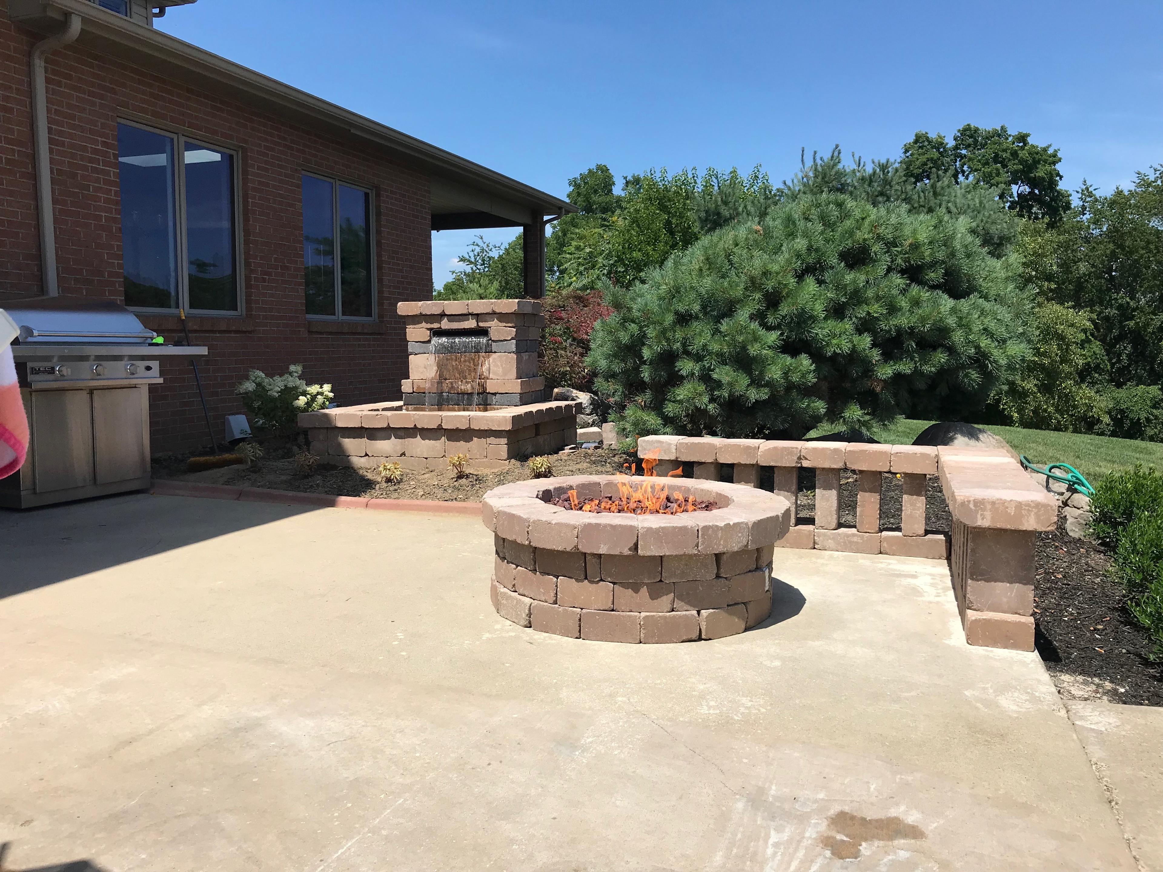 Concrete patio with round fire pit, stacked-stone waterfall feature, low wall, and grill area.