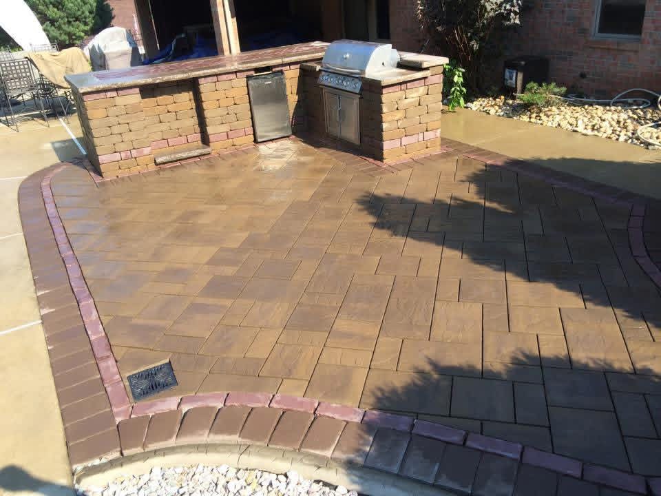 Brown paver patio with brick border and built-in stone outdoor kitchen with grill and refrigerator.