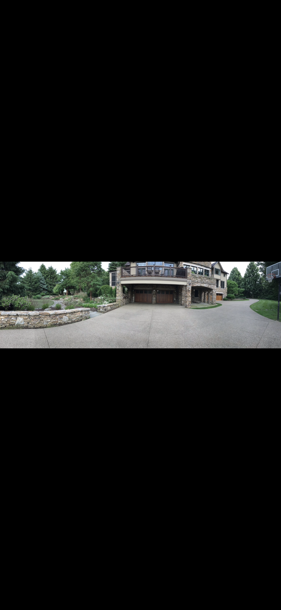 Panoramic view of a large stone-front home with a wide concrete drive, retaining wall, and professional landscaping.