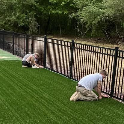 Two crew members installing a black metal picket fence along a green lawn beside a wooded area.