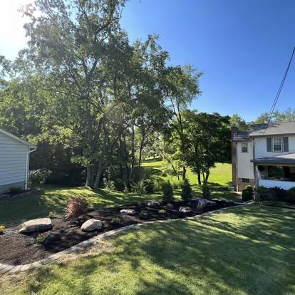 Curved garden bed with fresh mulch, decorative rocks, and a red Japanese maple on a green hillside.