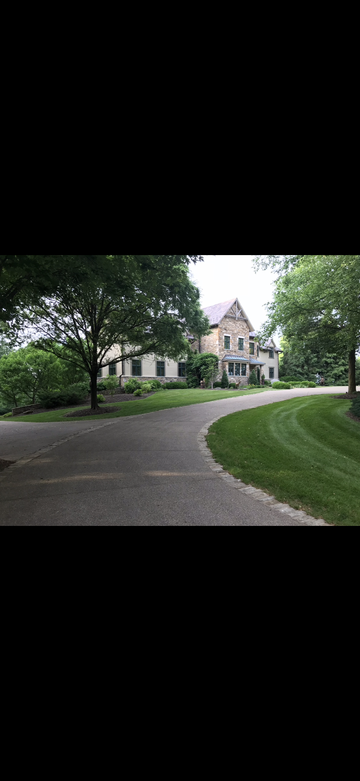 Two-story stone and siding home at the end of a curved driveway lined with lawn and mature trees.